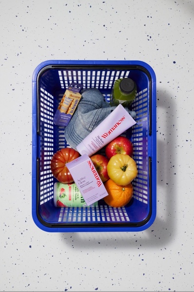 A top-down, flat-lay composition placing body cream and supplements inside a grocery basket alongside fresh fruit and vegetables.